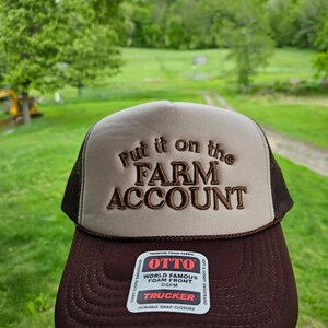 K&ouml;nnte beinhalten: Braune und beige Trucker-Cap mit dem gestickten Schriftzug "Put it on the FARM ACCOUNT". Die Kappe hat einen braunen Schirm und Mesh-Seiten. Das Etikett lautet "OTTO TRUCKER".