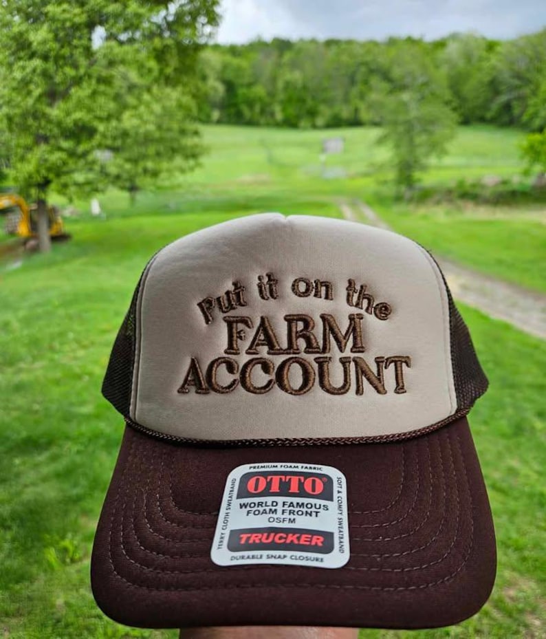 K&ouml;nnte beinhalten: Braune und beige Trucker-Cap mit dem gestickten Schriftzug "Put it on the FARM ACCOUNT". Die Kappe hat einen braunen Schirm und Mesh-Seiten. Das Etikett lautet "OTTO TRUCKER".