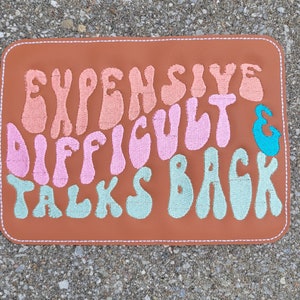 May include: A brown leather patch with the text "Expensive, Difficult & Talks Back" embroidered in pink, peach, and teal.
