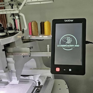 May include: A white Brother embroidery machine with a black touchscreen displaying the logo "LK Embroidery and Designs LLC." The machine has several spools of thread in shades of pink, brown, and yellow.