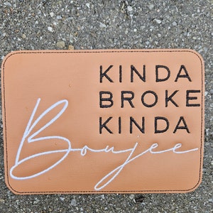 May include: A rectangular, tan-coloured patch with black stitched edges. The patch features the embroidered text "KINDA BROKE KINDA Boujee" in black and white. The background is a solid tan colour.