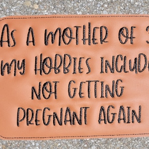 May include: A brown leather patch with black embroidered text that reads "As a mother of 3 my hobbies include not getting pregnant again".