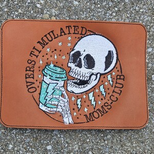 May include: Brown leather patch with embroidered design of a skull drinking from a coffee cup. The text around the skull reads "Overstimulated Moms Club".