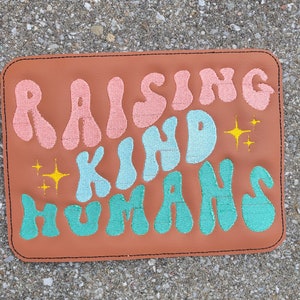 May include: A brown leather patch with the text "Raising Kind Humans" in pink, blue, and green lettering. The text is in a retro font style with yellow stars.