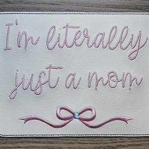 May include: White leather coaster with pink embroidery that reads "I'm literally just a mom" with a pink bow with a blue center.