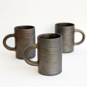 Pode incluir: Três canecas de cerâmica cinza escuro com alças arredondadas são dispostas sobre um fundo branco. As canecas têm formato cilíndrico e superfície texturizada. As canecas são adequadas para café, chá ou outras bebidas.