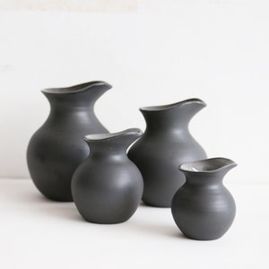 May include: Four matte black ceramic vases of different sizes are displayed. Each vase has a gently curved rim. The vases have a smooth, rounded shape, creating a minimalist aesthetic. The vases are measured in centimetres.