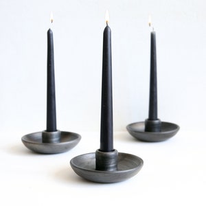 Könnte beinhalten: Drei schwarze Spitzkerzen mit brennenden Flammen werden in grauen Keramik-Kerzenhaltern präsentiert. Die Kerzenhalter haben ein flaches Schalendesign. Der Hintergrund ist eine schlichte weiße Oberfläche.