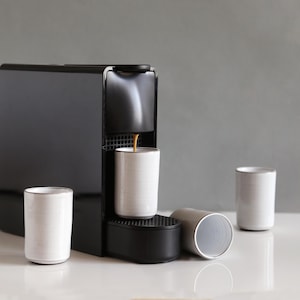 May include: A black espresso machine with a white ceramic cup being filled. Three additional white cups with silver rims are nearby. A clear glass container is to the left of the machine. The scene is set on a white surface against a gray background.