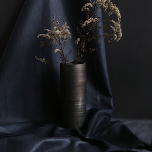 May include: A dark, cylindrical vase holds dried flowers against a dark blue backdrop. The vase has a textured, metallic appearance. The flowers are beige and add a natural element to the composition. The fabric drapes around the vase.