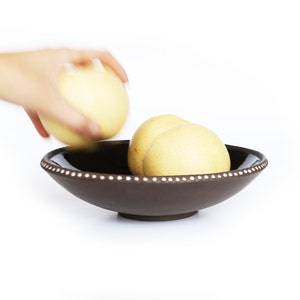 May include: A dark brown ceramic bowl with a white dotted rim contains several yellow fruits. A hand is reaching for another fruit, suggesting serving. The bowl is set against a white background.