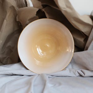 May include: A ceramic bowl with a gradient colour scheme, transitioning from a light yellow centre to a white rim. The bowl is set against a backdrop of crumpled brown paper and a grey fabric.