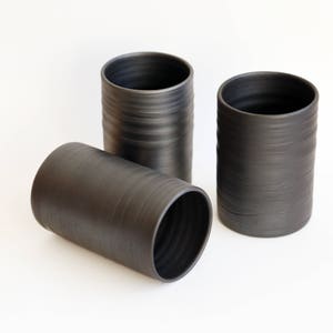May include: Three dark gray ceramic cylinders with textured surfaces. Two stand upright, while the third lies on its side. The cylinders have a simple, modern design, suitable for various decorative uses.