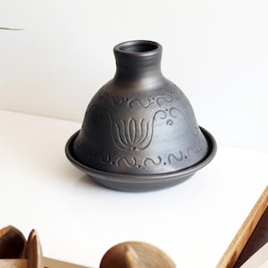 Może przedstawiać: Ciemnoszary ceramiczny garnek do tagine z pokrywką, z kwiatowym i wirującym wzorem. Garnek stoi na pasującej podstawie. Tagine to tradycyjne naczynie do gotowania.