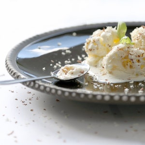 May include: Three scoops of vanilla ice cream on a dark grey plate, garnished with chocolate shavings and a sprig of mint. A spoon rests on the plate, filled with ice cream. The plate has a decorative beaded edge.