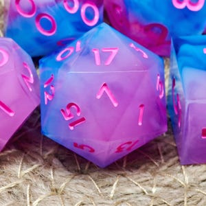 Op de afbeelding: Close-up van verschillende polyhedrale dobbelstenen in blauw- en paarstinten met roze nummering. De dobbelstenen zijn gemaakt van een doorschijnend materiaal, met een gemarmerd effect. De dobbelstenen zijn voor tabletop games.