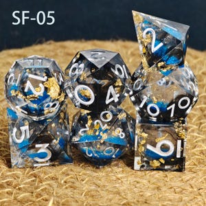 May include: A set of seven polyhedral dice with a clear, black, and blue resin design. The dice have gold leaf accents and white numbering. The text "SF-05" is in the upper left corner. These dice are for tabletop gaming.