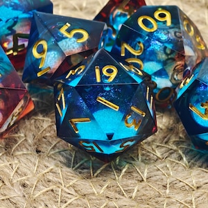 Op de afbeelding: Een verzameling veelvlakkige dobbelstenen met een hemels thema. De dobbelstenen zijn doorschijnend blauw met gouden cijfers en interne kleurwervelingen. De dobbelstenen zijn gerangschikt op een getextureerd, neutraal getint oppervlak.