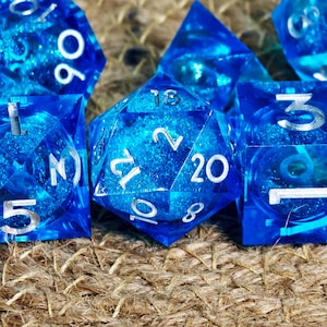Puede incluir: Primer plano de varios dados poliédricos azules con números blancos. Los dados están hechos de un material translúcido con un efecto arremolinado y veteado. Los dados están dispuestos sobre una superficie texturizada y marrón.