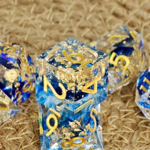 Op de afbeelding: Close-up van verschillende polyhedrale dobbelstenen. De dobbelstenen zijn helder met blauwe en gouden accenten. De cijfers zijn goudkleurig. De dobbelstenen zijn voor tabletop games.