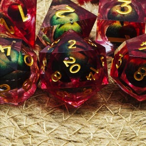May include: A collection of polyhedral dice with a red and gold color scheme. The dice have gold numbers and are made of a translucent material, revealing a green and black design inside. The dice are for tabletop games.