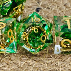 May include: Set of polyhedral dice with a green and gold color scheme. The dice are transparent with green swirls and gold numbering. The dice are designed for tabletop role-playing games.