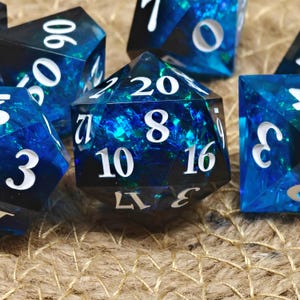 Può includere: Primo piano di diversi dadi poliedrici. I dadi sono blu scuro con un effetto scintillante simile a una galassia. I numeri bianchi sono visibili su ogni lato. I dadi sono per giochi da tavolo.