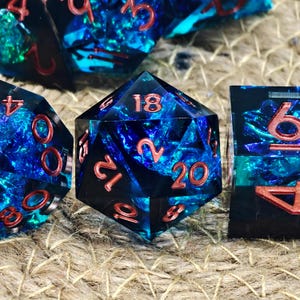 May include: Close-up of a set of polyhedral dice. The dice are a mix of black and blue with a shimmering, iridescent effect. The numbers are a reddish-orange colour. The dice are on a textured, tan surface.