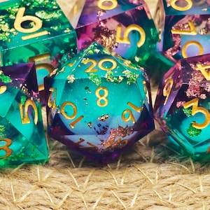 Peut inclure: Gros plan sur une collection de dés polyédriques colorés. Les dés sont turquoise, violets et dorés avec des chiffres dorés. Certains dés ont des accents de feuilles d'or. Les dés sont disposés sur une surface tissée.