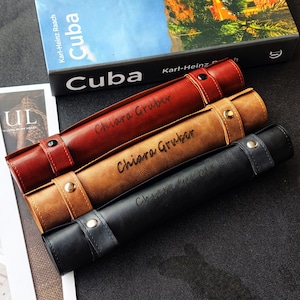 Könnte beinhalten: Drei Leder-Stifteetuis in Rot, Braun und Schwarz, jeweils mit Riemenverschluss und dem geprägten Namen "Chiara Gruber". Die Etuis sind zylindrisch und liegen auf einer dunklen Oberfläche, mit einem Buch mit dem Titel "Cuba" im Hintergrund.