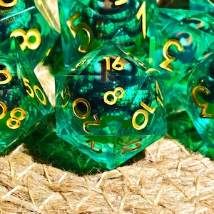 May include: A collection of translucent green polyhedral dice with gold-colored numbers. The dice have an internal design with a scale-like pattern. The dice are arranged on a textured, light brown surface.
