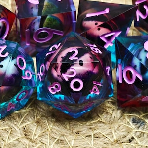 Puede incluir: Primer plano de un conjunto de dados poliédricos. Los dados son transparentes con una combinación de colores azul y morado. Los números son de color rosa brillante. Los dados están sobre una superficie texturizada de color beige.