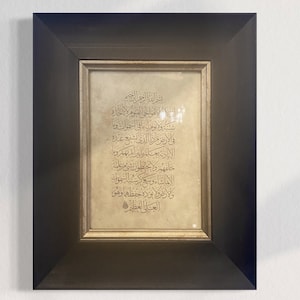 May include: A framed piece of art with a gold background and black frame. The art features Arabic calligraphy in a traditional style.