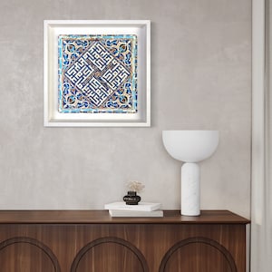 Peut inclure: Une image encadr&eacute;e blanche avec un motif de carrelage bleu et blanc. Le motif du carrelage pr&eacute;sente des motifs g&eacute;om&eacute;triques complexes et de la calligraphie arabe. L'image est accroch&eacute;e &agrave; un mur gris clair au-dessus d'un meuble en bois avec une lampe blanche.