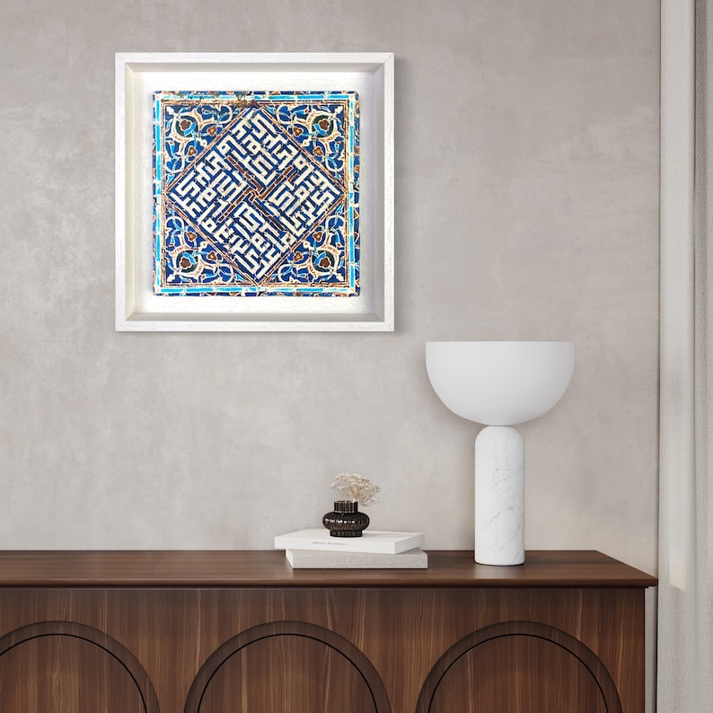 Peut inclure: Une image encadr&eacute;e blanche avec un motif de carrelage bleu et blanc. Le motif du carrelage pr&eacute;sente des motifs g&eacute;om&eacute;triques complexes et de la calligraphie arabe. L'image est accroch&eacute;e &agrave; un mur gris clair au-dessus d'un meuble en bois avec une lampe blanche.