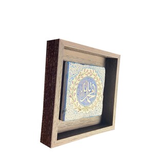 May include: A framed, square, blue and white tile with Arabic calligraphy. The tile is decorated with floral patterns in pink and green. The frame is made of dark wood.