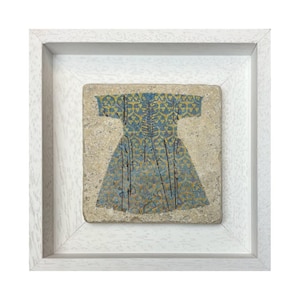 Op de afbeelding: Een ingelijst kunstwerk met een blauw en geel patroon van een jurk. Het kunstwerk staat tegen een getextureerde, beige achtergrond en is ingekaderd in een witte, vierkante lijst. Het jurk-ontwerp heeft ingewikkelde details.