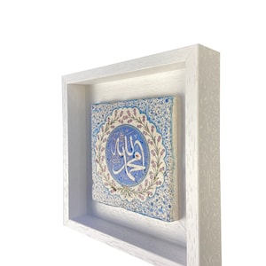 May include: A square, framed ceramic tile with an Islamic design. The tile features a central blue medallion with Arabic calligraphy, surrounded by floral patterns in blue, pink, and white. The frame is a simple, white wooden box.
