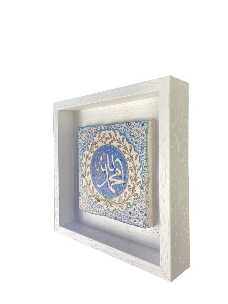 May include: A square, framed ceramic tile with an Islamic design. The tile features a central blue medallion with Arabic calligraphy, surrounded by floral patterns in blue, pink, and white. The frame is a simple, white wooden box.