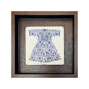 Op de afbeelding: Een ingelijst kunstwerk met een blauw-wit patroon van een kledingstuk. Het kunstwerk is geplaatst in een donkerbruine houten lijst, wat een contrast creëert met de lichtgekleurde achtergrond van het kunstwerk.