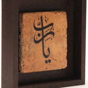 May include: A framed piece of art with Arabic calligraphy on a terracotta tile. The tile is a warm orange color and the calligraphy is in black ink. The frame is dark brown wood.