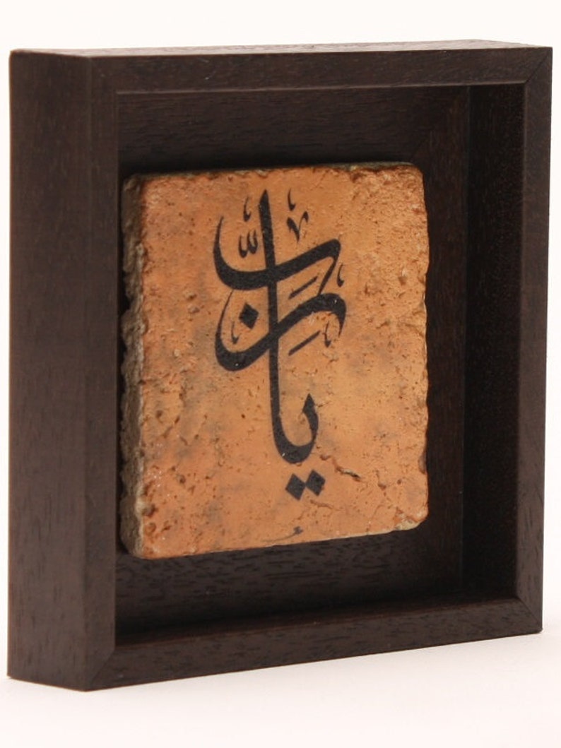 May include: A framed piece of art with Arabic calligraphy on a terracotta tile. The tile is a warm orange color and the calligraphy is in black ink. The frame is dark brown wood.