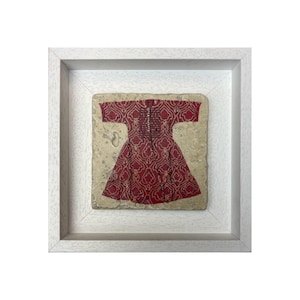 Op de afbeelding: Een ingelijst kunstwerk met een rood patroon van een jurk. De jurk staat tegen een beige achtergrond en is ingelijst in een witte vierkante lijst. De jurk heeft een gedetailleerd patroon en is gecentreerd in de lijst.