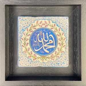 May include: Square framed artwork featuring Islamic calligraphy in a blue and white circular design. The artwork is set against a cream-colored background with floral patterns and framed in a black wooden frame. The calligraphy is in a script style.