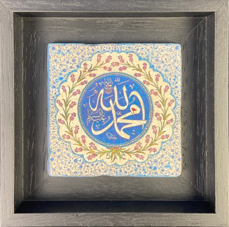 May include: Square framed artwork featuring Islamic calligraphy in a blue and white circular design. The artwork is set against a cream-colored background with floral patterns and framed in a black wooden frame. The calligraphy is in a script style.
