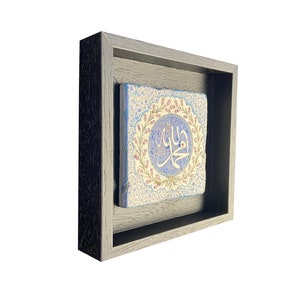 May include: A framed, square ceramic tile featuring Arabic calligraphy in a light blue circle with a floral border. The tile is set within a dark gray wooden frame, creating a layered effect. The background is white.