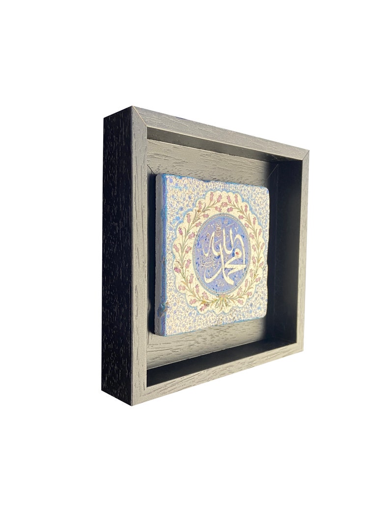 May include: A framed, square ceramic tile featuring Arabic calligraphy in a light blue circle with a floral border. The tile is set within a dark gray wooden frame, creating a layered effect. The background is white.