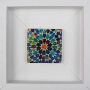 Op de afbeelding: Een vierkante, kleurrijke tegel met een geometrisch patroon, omlijst in een witte houten schaduwdoos. De tegel heeft een centraal sterrenpatroon in tinten oranje, rood, groen, blauw en blauwgroen. De lijst is een eenvoudig, strak ontwerp.