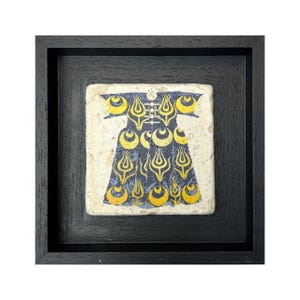 Op de afbeelding: Ingeklemd kunstwerk met een gestileerd ontwerp in marineblauw en geel op een getextureerde, gebroken witte achtergrond. Het ontwerp, dat op een gewaad lijkt, is gevat in een zwarte vierkante lijst. Het kunstwerk is vierkant.