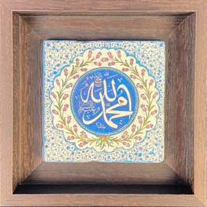 May include: Square ceramic tile with Arabic calligraphy in a blue circle, surrounded by a floral design. The tile is set in a dark brown wooden frame. The background has a light blue and white floral pattern.